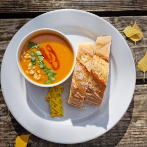 Thai soup with crusty bread and butter in white bowl on white plate on wooden picnic table